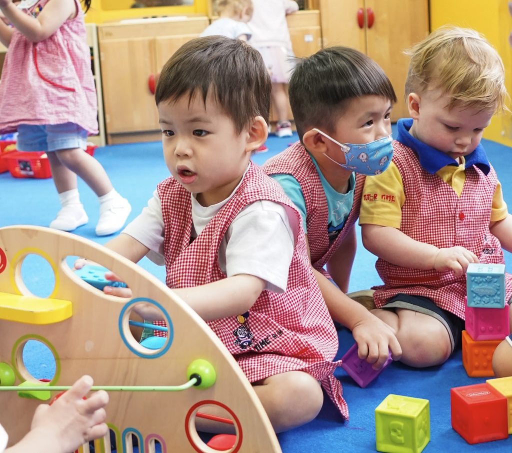 Toddlers in red aprons play with toys in a vibrant classroom; one with a bead maze, another stacking blocks.